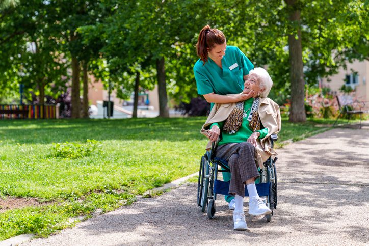 An elderly woman in a wheelchair is tenderly caressed by her caring nurse as they take a sunny stroll through a lush, tree-filled park in the city