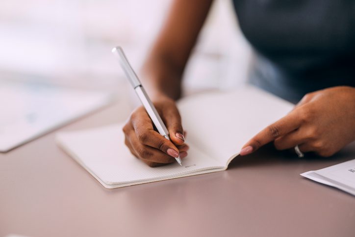 A professional person writing in a notebook at a desk, highlighting focus and organization in a business setting. The image emphasizes planning, note-taking, and productivity.