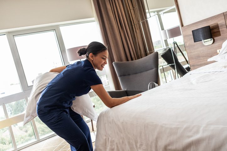 A female hotel staff member is preparing a room for the next guests. She carefully makes the bed, arranges pillows, and ensures the room is tidy and ready for service.