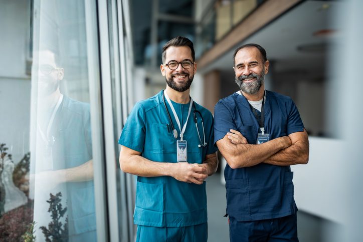 Two male doctors and coworkers smiling, wearing scrubs and stethoscopes, standing in a bright hospital corridor, symbolizing medical partnership and healthcare professionalism